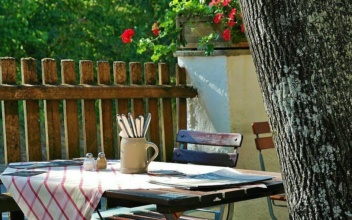 In einem Biergarten steht ein eingedeckter Tisch hinter einem Baum. In einem Biergarten steht ein eingedeckter Tisch hinter einem Baum.